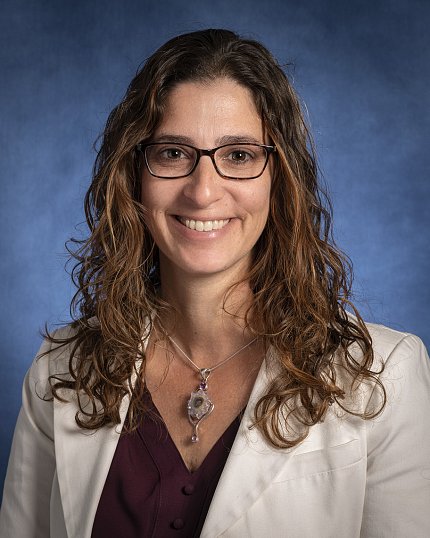 Headshot of D'Souza against a dark blue background. She is wearing glasses and a white jacket.