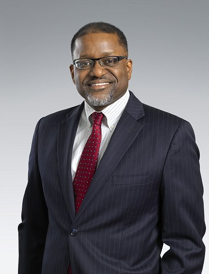 Gibbons, wearing a suit and tie, standing against a light gray background.