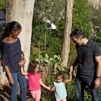 Two parents hold the hands of their young children as they navigate a forested trail.