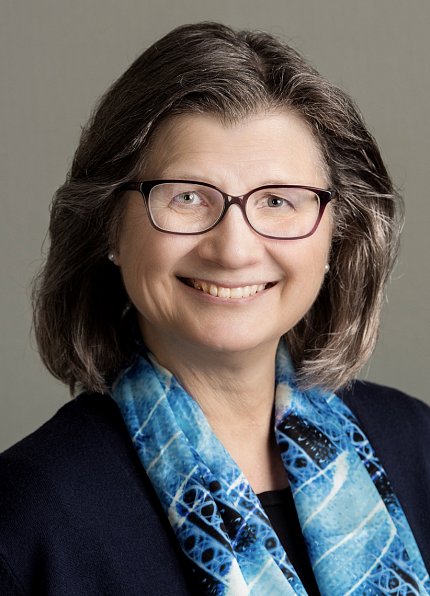 Headshot of Giger, who wears glasses, a blue scarf and black jacket, and smiles against a gray backdrop.