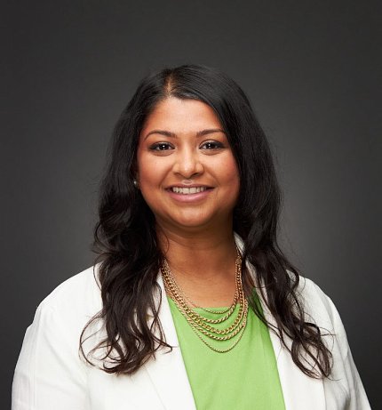 Mistry Gosine, wearing layered gold necklaces over a green shirt and white lab coat, smiles against a dark gray backdrop