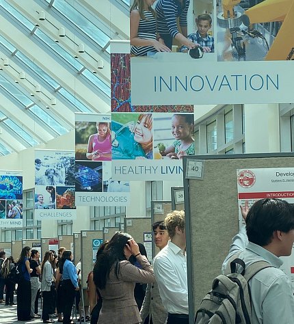 Rows of scientific posters in a large atrium. People cluster around the posters to listen to student presentations.