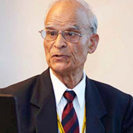 A bespectacled Pant speaks to someone outside of the frame. He wears a black suit and red and black striped tie.