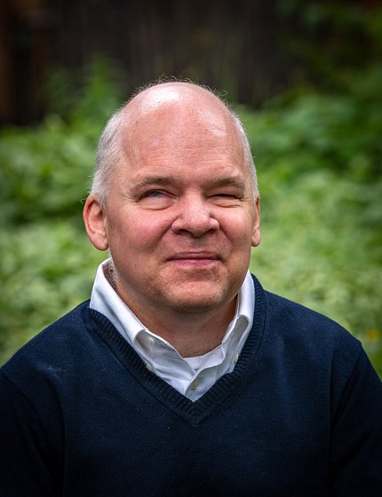Weed, wearing a white collared shirt and navy pullover, smiles against a background of green foliage.