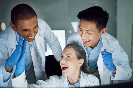 Three smiling scientists in lab coats high-five each other in front of a computer.