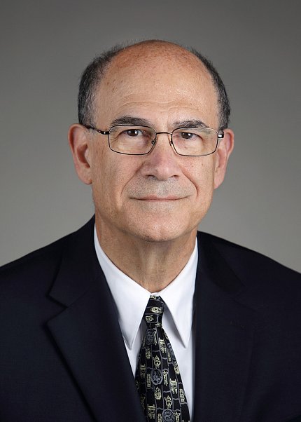 Headshot of Hallett, who is wearing a suit, dark patterned tie, and glasses and pictured against a light gray background.