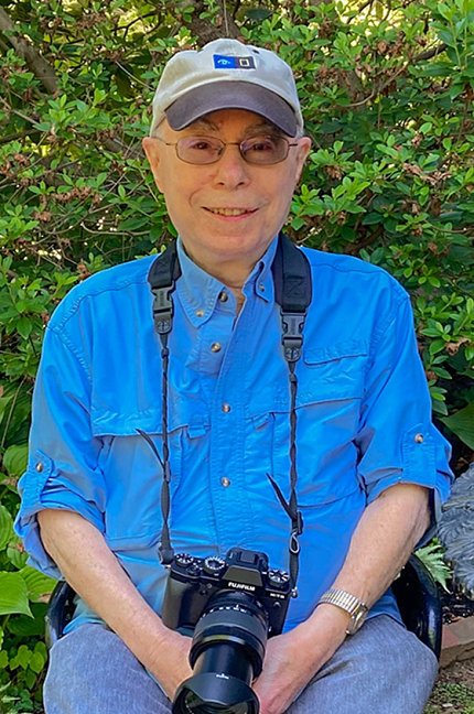 Jacobson sits on a chair in front of green foliage. He is wearing a blue shirt and a ball cap and holds a camera in his lap.