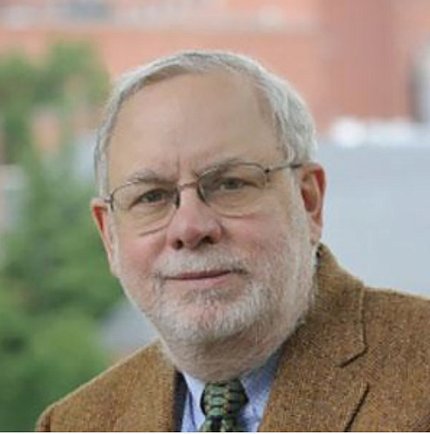 Photo of a bespectacled Dr. Schechter, who is wearing a brown jacket and a tie. He is pictured outdoors with a blurry tree and building in the background.