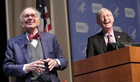 Dr. Kandel smiles on stage as a grinning Dr. Collins looks toward audience in Masur auditorium