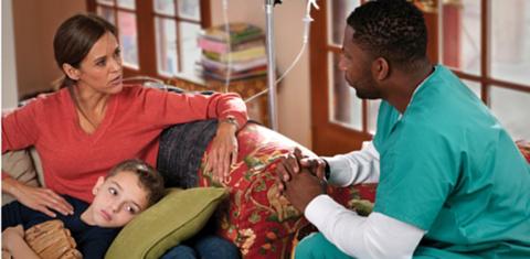 A mom, while son lays on her lap, talks with a doctor in her home.