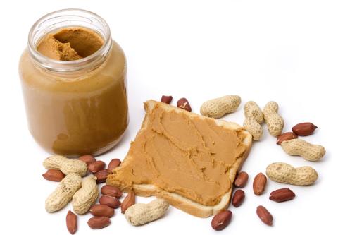A jar of peanut butter sits on table next to slice of bread slathered with peanut butter as well as peanuts and shells.