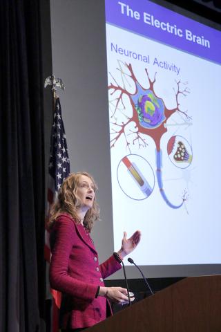Lisanby speaks from behind the microphone with slide projected beside her