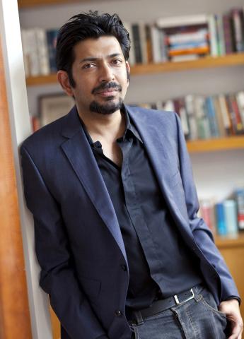 Mukherjee leans against a wall in front of a bookshelf.