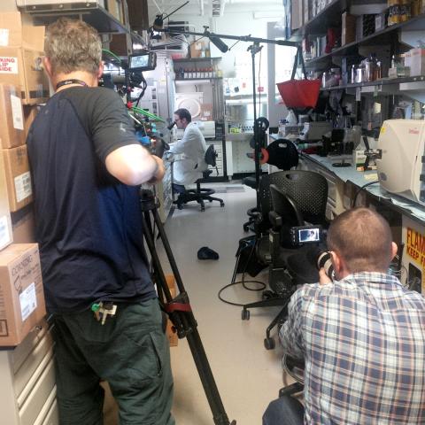 Man seated in lab coat at microscope as seen from behind two cameramen