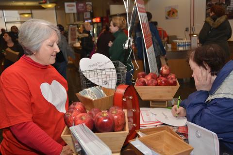 Woman in red heart sweatshirt stands behind table display of red apples as visitor looks on.