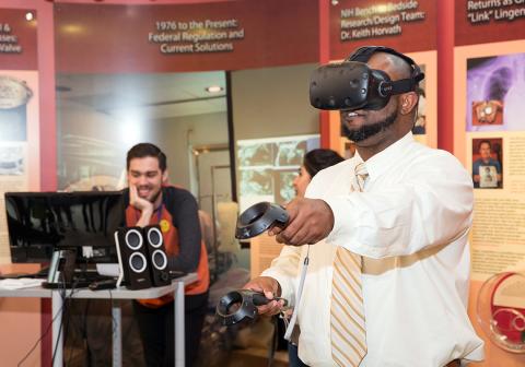 A man holds out, moves handheld device and wears head gear to try virtual reality experience in Clinical Center south lobby.