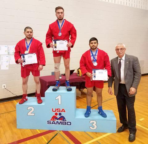 Three athletes wearing uniforms and medals on various levels of portable podium, alongside a coach