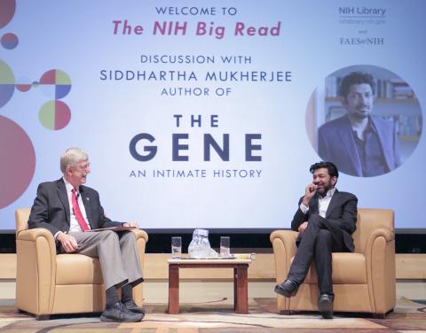 Collins and Mukherjee seated on stage with slide announcing event projected behind them. 