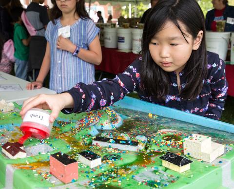 At Earth Day event outside, 7-year-old Hanna Cho examines green model of a town, pouring beads representing trash from a container to learn effects of pollution.