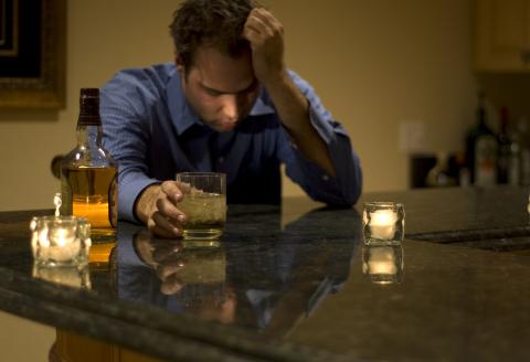 A man sits at kitchen counter, forlorn, holding his head in one hand and a glass of whiskey in the other