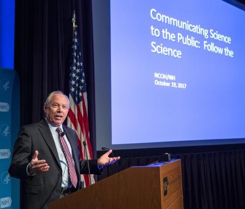 Leshner speaks with hands open at podium, next to slide that reads: Communicating Science to the Public