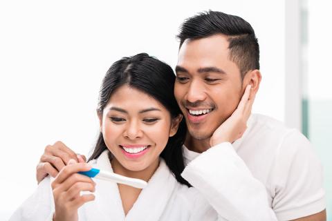 Woman smiling, holding pregnancy test results with man smiling over her shoulder 