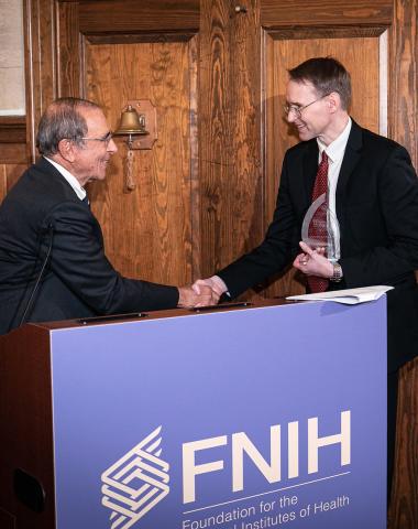 Gallin shakes hands with awardee Kochenderfer at podium draped with purple FNIH banner.