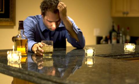 Man seated at bar, one hand holding a glass with alcoholic beverage, other hand against his bowed head