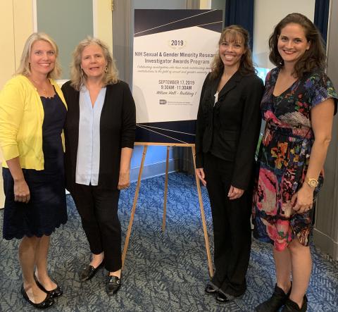 The 3 smiling honorees stand with director of the Sexual and Gender Minority Research Office in front of Investigator Awards Program poster.