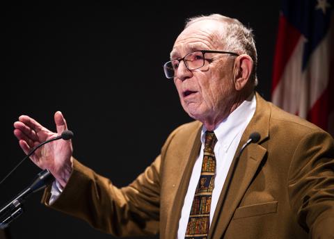 Feigenbaum stands on stage with outstretched hand.