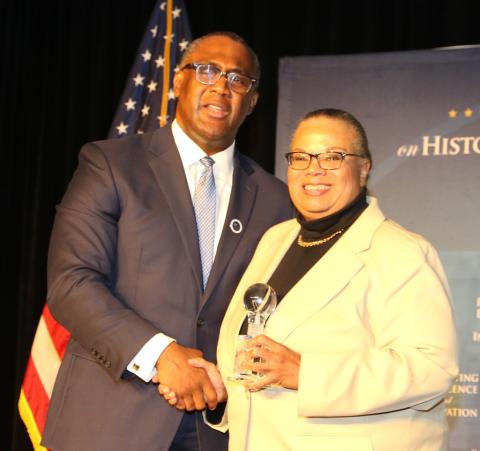 A smiling Diane Frasier shakes hands with Johnathan Holifield on stage in front of American flag.