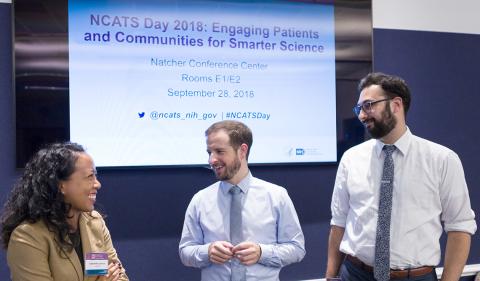 Three participants in NCATS Day 2008 gather in front of projected slide announcing the event.