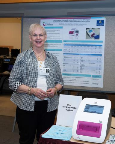 Gaydos stands beside diagnostic instrument, with scientific poster behind her.