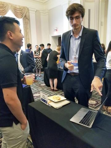 Sengul behind table, gestures toward open laptop.