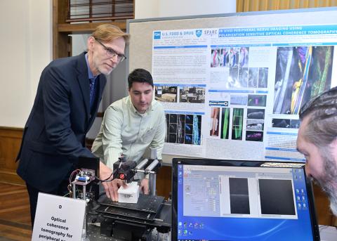 Tromberg and Monroy manipulate an imaging device as Hammer views screen, with scientific poster behind them. 