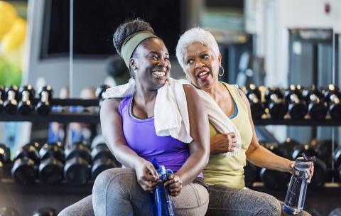 Two women sit smiling with water bottles in front of hand weights at a gym.