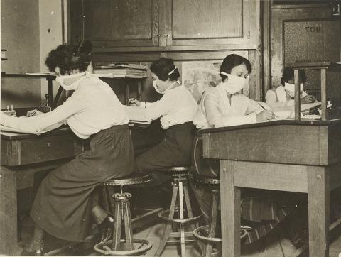 A black &amp; white photo shows four women wearing masks working at their desks.