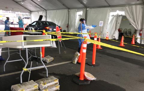 Outside, under a tent, responders in gowns, masks and gloves stand ready to test people for covid at a drive-through testing facility.