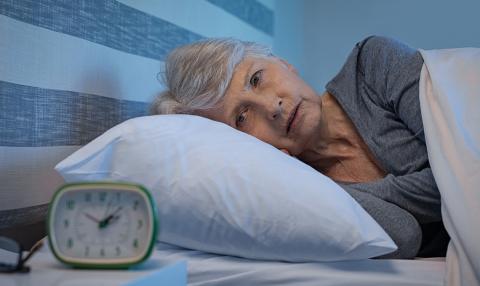 An older woman lies awake in bed next to an alarm clock on her nightstand.