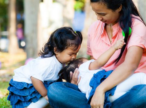 A mom sits on the lawn snuggling with her 2 young children.
