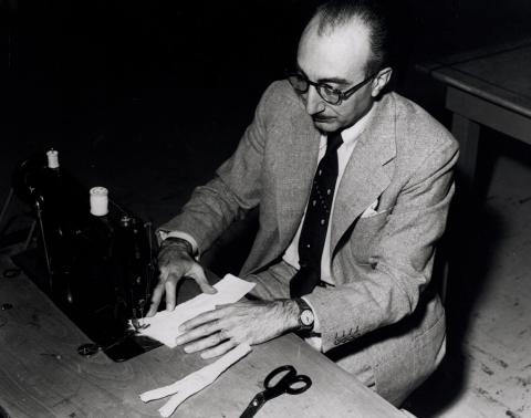 Archival photo shows DeBakey stitching strips of Dacron using a sewing machine, with scissors on table next to him