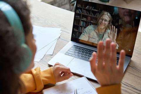 A woman sits at her desk with her laptop; she and her instructor on screen are waving to each other.