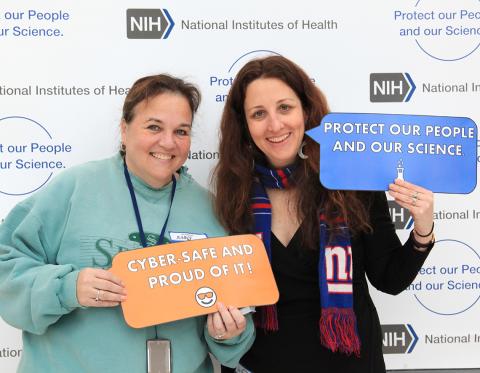 Two NIH'ers hold signs: "protect our people and our science" and "cyber safe and proud of it."