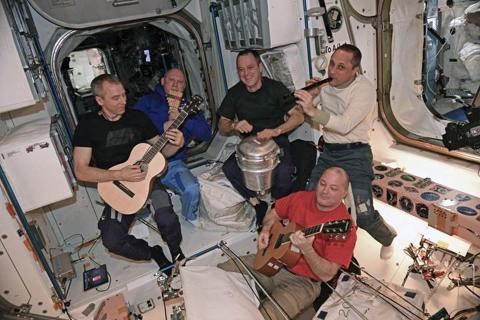 Astronauts play musical instruments, including a “drum” consisting of a repurposed solid waste container.