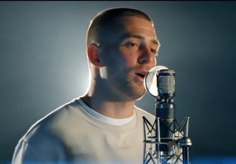 Thompson, in white shirt, sings into a microphone with spotlight behind him.