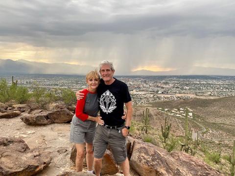 Atop a large rock, Warren and Beiser smile toward camera, with a stormy sky and a cityscape behind them.