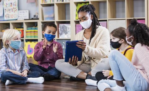 A teacher is seated holding up a tablet for 4 students seated around her, all wearing masks.