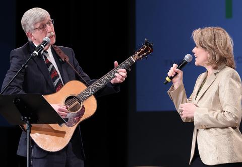 Collins holding guitar and Fleming holding a microphone sing together on stage in Masur Auditorium in 2019.