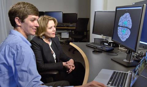 A researcher and Fleming sit in front of a computer screen showing an image of her brain with different colors reflecting when her brain was activated during her MRI.
