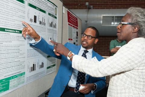 Gesturing toward a poster, black male scientist talks with black female scientist.